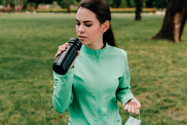 Ελκυστική αθλήτρια κρατώντας αθλητικό μπουκάλι και ιατρική μάσκα στο πάρκο  - Φωτογραφία, εικόνα