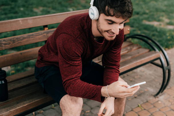 Glimlachende sportman met smartphone en koptelefoon op bankje in park  - Foto, afbeelding