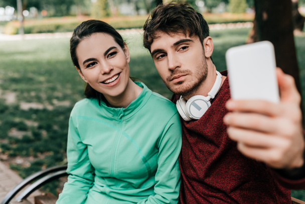 Parktaki bankta oturan yakışıklı erkek arkadaşın yanında akıllı telefonuyla selfie çeken gülümseyen sporcu kadının seçici odağı.  - Fotoğraf, Görsel