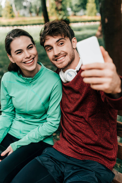 Επιλεκτική εστίαση του χαμογελαστού αθλητή λήψη selfie με smartphone κοντά φίλη στον πάγκο στο πάρκο  - Φωτογραφία, εικόνα
