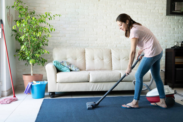 Profile view of a beautiful Caucasian woman using a vacuum cleaner on a carpet at home and looking happy - Photo, Image