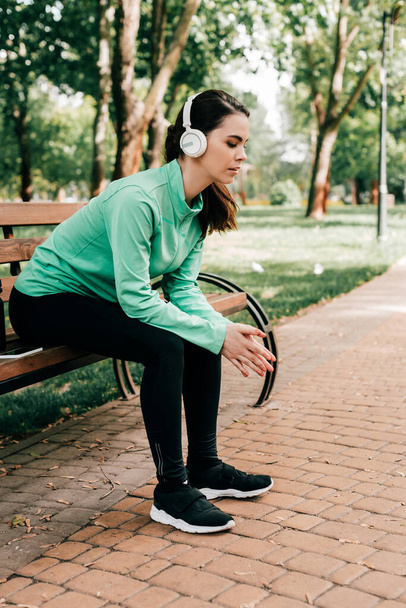 Side view of young sportswoman in headphones resting on bench in park  - Fotó, kép