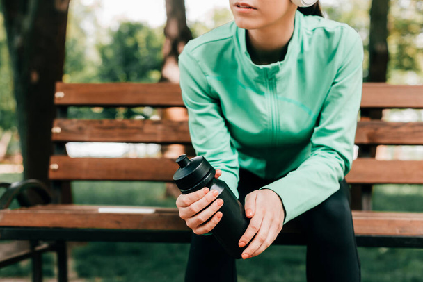 Cropped view of sportswoman holding sports bottle while sitting on bench in park  - Foto, imagen