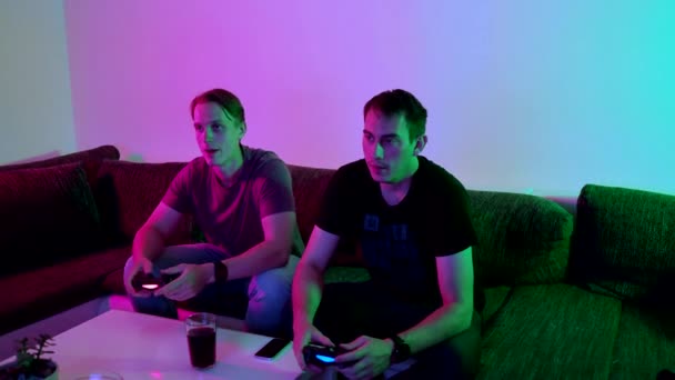 Two happy friends playing action video game in the living room sitting on the sofa in 4K VIDEO. Intense competition between two young male players is full of emotions.  - Кадры, видео
