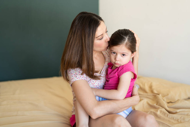 Retrato de una niña molesta siendo consolada por su madre mientras está sentada en una cama en casa
 - Foto, Imagen