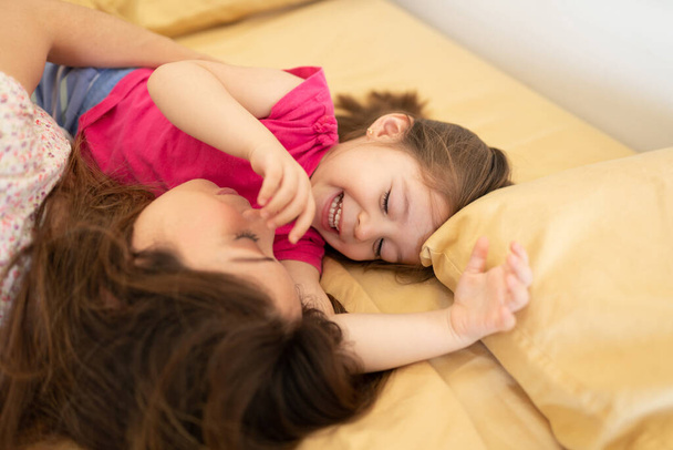Glückliches kleines kaukasisches Mädchen, das seine Mutter weckt und lächelt, während es sich zu Hause ein Bett teilt - Foto, Bild