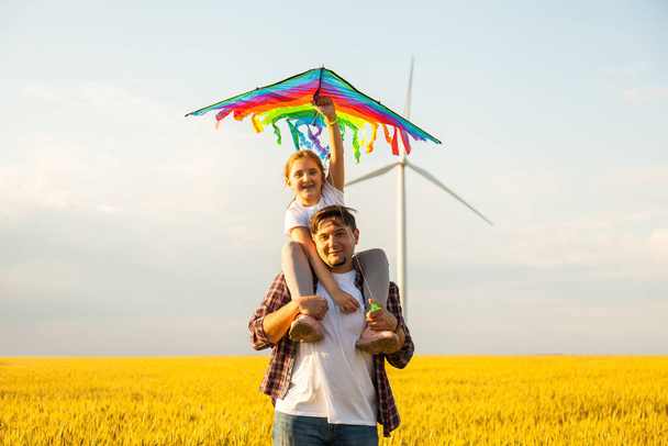 Ευτυχισμένος πατέρας δίνει στην κόρη της μια βόλτα με την πλάτη, διασκεδάζοντας, παίζοντας με χαρταετό μαζί στο χωράφι με το σιτάρι - Φωτογραφία, εικόνα