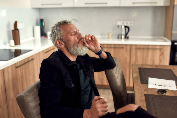 Breathe in. Middle-aged man smoking a marijuana cigarette or joint, sitting at home, in the kitchen. Marijuana grinder and weed on the table - Photo, image