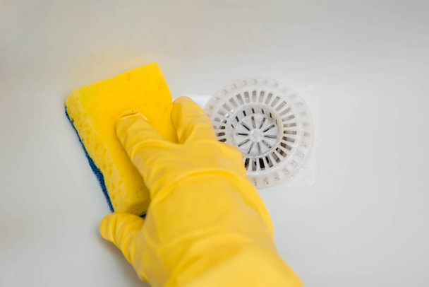 Close-up van een hand in een gele rubberen handschoen veegt een witte gootsteen af met een spons. - Foto, afbeelding