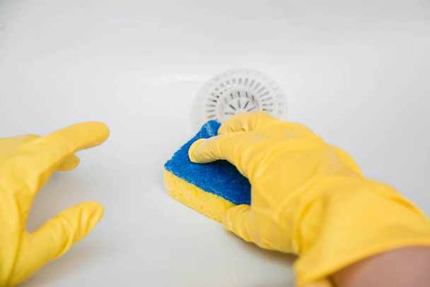 Close-up of a hand in a yellow rubber glove wipes a white sink with a sponge. - Foto, afbeelding