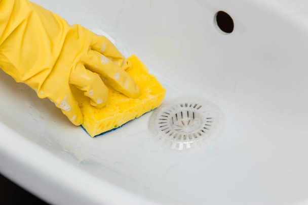 Primer plano de una mano en un guante de goma amarillo limpia un fregadero blanco con una esponja
. - Foto, imagen