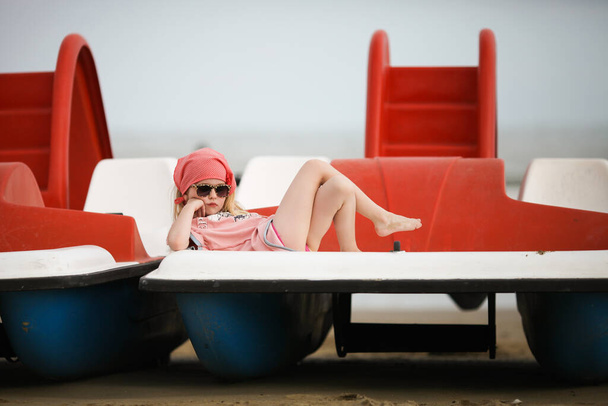 Dziewczynka w okularach leżała na łodzi na plaży. Smutna blondynka marzy o czymś na rowerze. Lido di Jesolo, Włochy. Wakacje z dziećmi koncepcja. - Zdjęcie, obraz