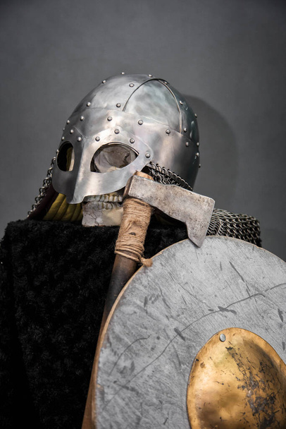 Skull in a Viking helmet, on a black pedestal, in front of him an ax and a round shield. Studio photo on a gray background. - Foto, Bild