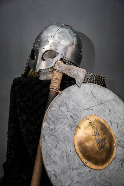 Cráneo en un casco vikingo, sobre un pedestal negro, delante de él un hacha y un escudo redondo. Foto del estudio sobre un fondo gris
. - Foto, Imagen