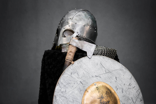 Skull in a Viking helmet, on a black pedestal, in front of him an ax and a round shield. Studio photo on a gray background. - Fotografie, Obrázek