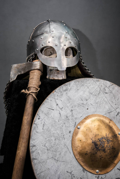 Skull in a Viking helmet, on a black pedestal, in front of him an ax and a round shield. Studio photo on a gray background. - Foto, Imagem