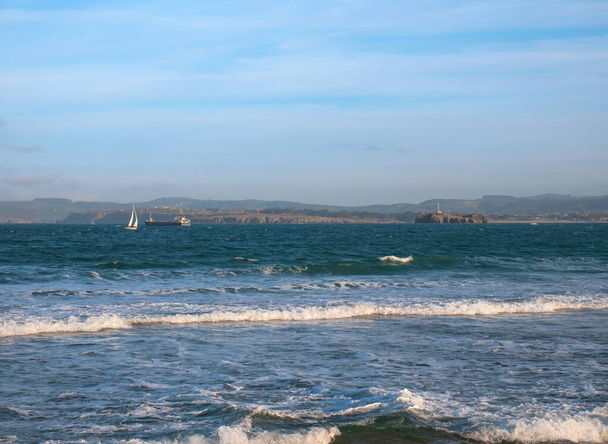 ein Öltanker und ein Segelboot fahren entlang des kantabrischen Meeres in der Nähe der Bucht von Santander mit dem Leuchtturm und der kantabrischen Küste im Hintergrund - Foto, Bild