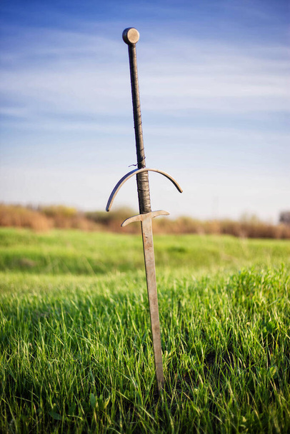 two-handed sword in the ground, against the background of a green field, sky and trees. Lost, forgotten sword, fallen warrior. - Fotografie, Obrázek