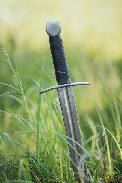 Knight's sword pierced to the ground. In the high yellow-green grass. The battlefield, after the battle. Crusade, medieval weapons. A bright sunny day in the steppe. - Foto, Bild