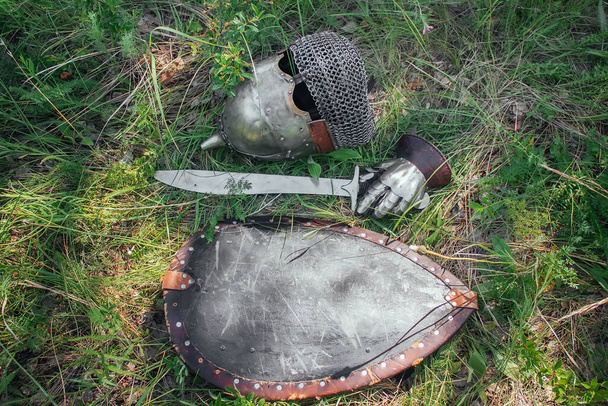 A helmet, a sword, a steel glove and a shield of a Mongolian warrior lie on the ground in a tall green grass. Lost, taken after the battle. Medieval weapons of a nomad, steppe soldier. - Valokuva, kuva