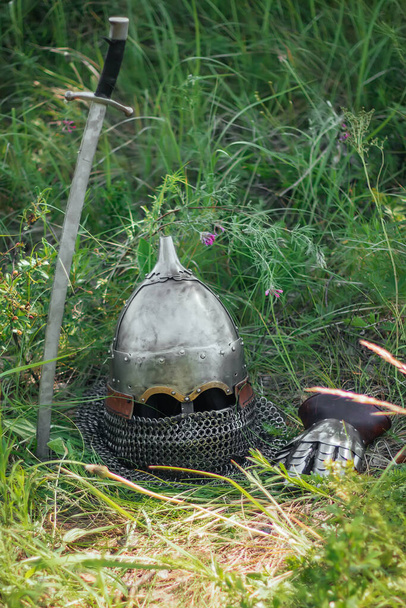 The helmet, sword and glove of a Mongolian warrior lie on the ground in a tall green grass. Lost, taken after the battle. Medieval weapons of a nomad, steppe soldier. - Foto, imagen