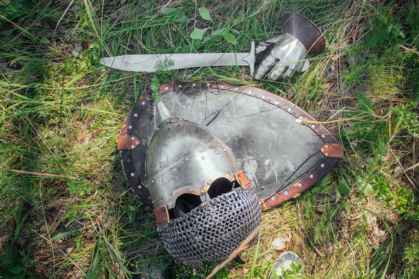 A helmet, a sword, a steel glove and a shield of a Mongolian warrior lie on the ground in a tall green grass. Lost, taken after the battle. Medieval weapons of a nomad, steppe soldier. - Foto, Bild