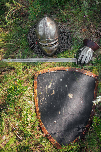 Helmet, sword, steel glove and triangular shield, lie on the ground in high green grass. The lost weapons after the battle, the battlefield. Medieval weapons of the knight. - Photo, Image