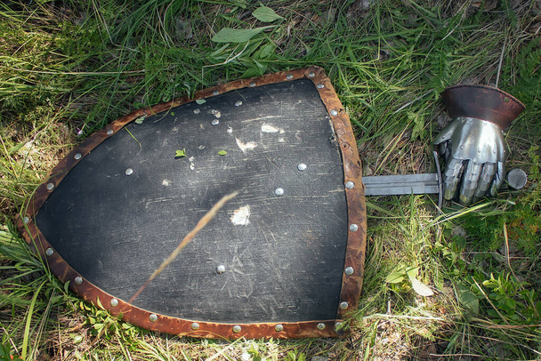 Sword, steel glove and triangular shield, lie on the ground in high green grass. The lost weapons after the battle, the battlefield. Medieval weapons of the knight. - Zdjęcie, obraz