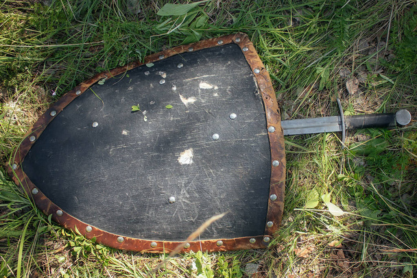 Sword lying on a triangular shield, on the ground in a tall green grass. The lost weapons after the battle, the battlefield. Medieval weapons of the knight. - Zdjęcie, obraz