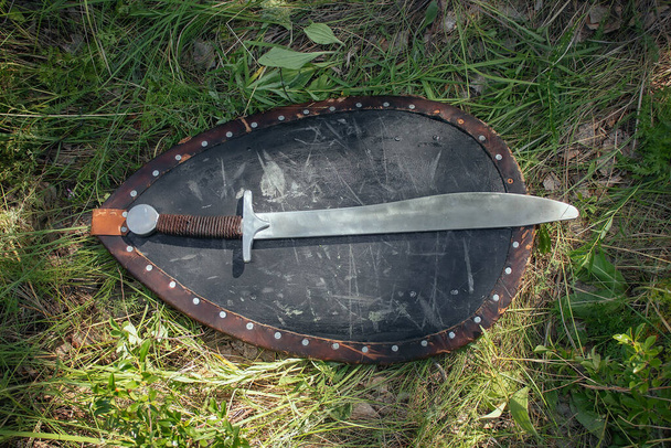 Saber and Shield of the Mongolian soldier lie on the ground in high green grass. Lost, taken after the battle. Medieval weapons of a nomad, steppe soldier. - Фото, изображение