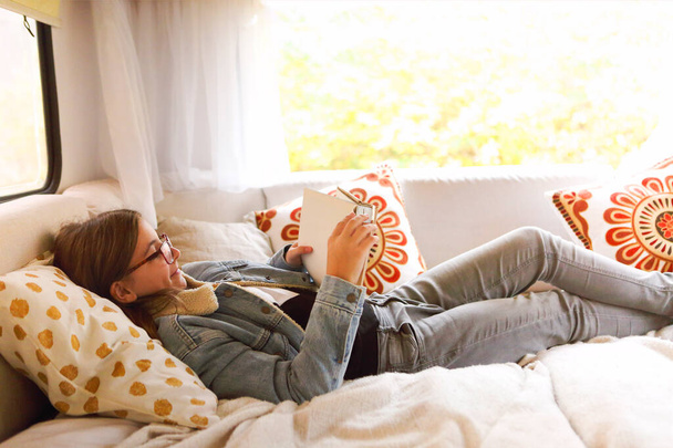 Widok z boku pozytywnej nastolatki w ubraniach casual i okulary leżące na łóżku w pobliżu okna i czytania książki podczas odpoczynku wewnątrz kampera - Zdjęcie, obraz