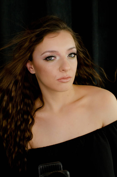 belle jeune fille brune regardant une caméra avec un maquillage professionnel, habillée en noir, avec des cheveux flottants, épaule nue, sur fond noir, plan studio, image verticale
 - Photo, image