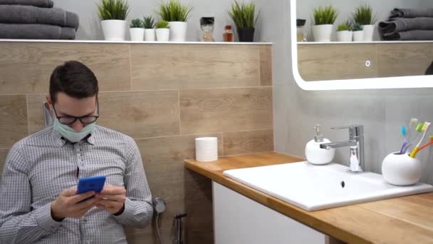 Young Caucasian nice man in gray plaid shirt, protective disposable face mask, black glasses sit in bathroom on toilet bowl, use phone, check news feed. Isolation morning at home during quarantine. Inside view. - Кадры, видео