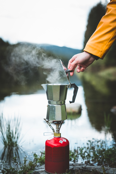 Menschliche Hand brüht Geysir-Kaffeemaschine, während sie kochenden Tee oder Kaffee auf dem Campinggasherd am See zubereitet. Camping Kochen in der Natur im Freien. Erholung im Freien und Lebensstil auf dem Campingplatz. - Foto, Bild