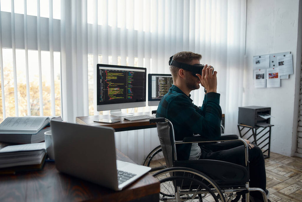 Young male web developer in a wheelchair wearing virtual reality glasses while working at his workplace in modern office. Multiple computer screens with program code - Valokuva, kuva