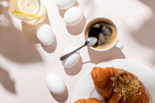 top view of chicken eggs, water with lemon, coffee and croissants for breakfast on grey table with shadows - Foto, afbeelding