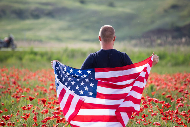 Attractive man holding Flag of the United States in beautiful poppy field on a clear, sunny day. Celebrating Independence Day, National holiday concept. - Photo, Image
