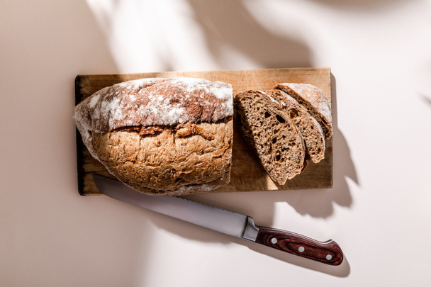 ylhäältä näkymä tuoretta paistettua ja viipaloitua leipää leikkuulaudalla veitsellä harmaalla pöydällä varjoilla - Valokuva, kuva