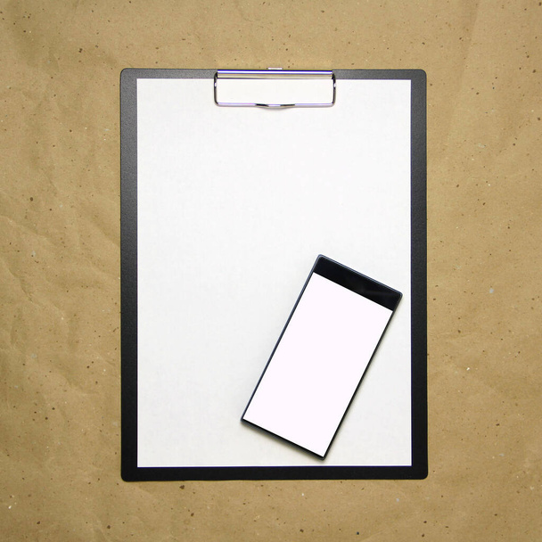The tablet with a clip for the clip of paper with a white sheet a4 lies against the background of craft brown crumpled paper. Above is a mobile phone with a white screen. Place for text and layout for design. - Photo, image