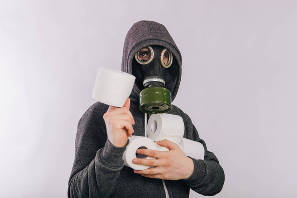Un tipo con una máscara de gas y una sudadera gris en una capucha sostiene rollos de papel higiénico en sus manos sobre un fondo blanco. El hedor en el baño. Pánico durante la cuarentena
. - Foto, Imagen