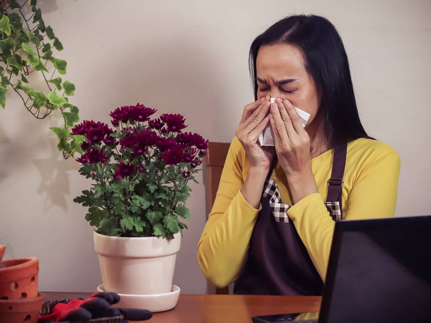 Aasialainen naispuutarhuri, jolla on allergia, istuu puupöydässä kukkien kanssa kukkaruukussa, pitää kasvokudosta ja aivastaa. - Valokuva, kuva