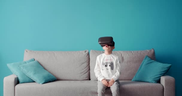 Happy little boy sitting on couch in virtual glasses - Materiał filmowy, wideo