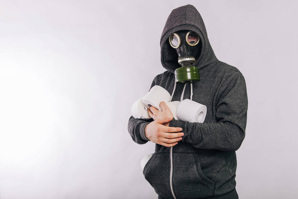 A guy in a gas mask and a gray sweatshirt in a hood holds rolls of toilet paper in his hands on a white background. The stink in the toilet. Panic during quarantine. - Photo, Image