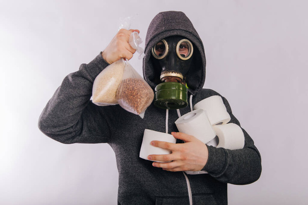 A guy in a gas mask and a gray sweatshirt holds a pack of buckwheat and paper. The stink in the toilet. Coronavirus pandemic - 写真・画像