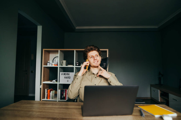 Pozitív fiatalember ingben otthon ül a munkahelyén egy laptopon, és mosollyal az arcán telefonál. Mosolygó szabadúszó dolgozik távolról otthonról a háttérben egy hangulatos hálószoba. - Fotó, kép