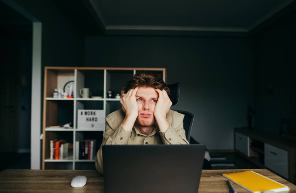 Väsynyt nuori mies paidassa istuu kotona kannettavan tietokoneen kanssa etätyössä, kädestä pitäen päänsä epätoivoisena ja katsoen ylös surullisella naamalla. Tyytymätön freelancer työskentelee kotona. - Valokuva, kuva