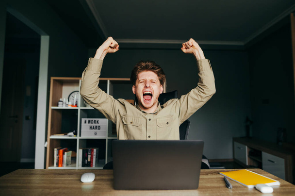 Joyeux pigiste dans une chemise est assis à un bureau avec un ordinateur portable et se réjouit émotionnellement avec ses mains en l'air. Portrait d'un jeune homme heureux au travail à distance se réjouit dans le succès.
. - Photo, image