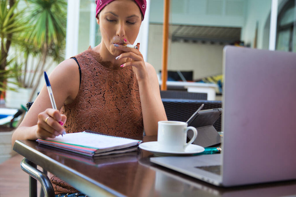 Kobieta z turbanem na głowie, pisząca w notesie, paląca papierosa na tarasie przed laptopem i kawą. - Zdjęcie, obraz