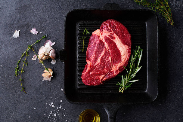 Black Angus prime beef chuck roll steak on cast iron grill pan with fresh rosemary and garlic with  olive oil and thyme. Creative layout with cooking ingredients, Top view    - Foto, immagini