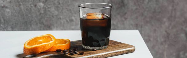 koude koffie zetten met ijs in glas in de buurt van sinaasappelschijfjes op snijplank en koffiebonen op witte tafel, panoramisch schot - Foto, afbeelding